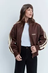 Cazadora de estilo deportivo en color marrón, confeccionada en tejido tipo gamuza. Presenta cierre frontal completo, cuello alto, puños y ruedo acanalados, y franjas laterales en tono rosado. Incluye el logo de la marca bordado en el pecho.