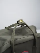 Mochila color verde militar con diseño clásico y minimalista. Cuenta con un compartimento principal con cierre de doble cremallera, un bolsillo frontal con cierre y dos asas superiores con broche. En el frente, tiene un parche circular con el texto "FORUM INDUSTRIES CO." en rojo.