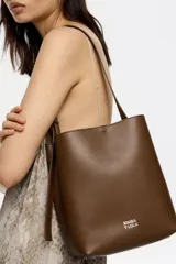 Bolso de hombro tipo cubo de estructura rígida, color marrón oscuro, confeccionado en piel vacuna. Posee asa regulable con hebilla metálica y cierre de cordón en la parte superior. Lleva el logo metálico de Bimba y Lola en la parte frontal inferior.