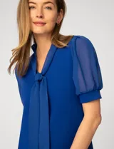 Blusa azul de corte recto con escote en V y lazo decorativo integrado en el cuello. Presenta mangas cortas abullonadas confeccionadas en gasa semitransparente con puños ajustados.