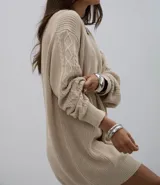 Vestido corto de punto en color beige, con diseño de trenzas en las mangas y cuello redondo. Presenta un corte holgado y puños acanalados.