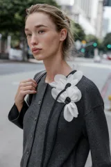 Chaqueta gris de punto con cuello redondo, manga larga y cierre frontal con botones. Detalle de flor blanca y negra en el frente. Tejido en rib.