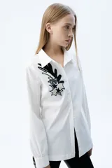 Camisa de corte recto con cuello clásico y cierre frontal de botones. Presenta un parche bordado con mostacillas en tonos blanco y negro en el frente, con diseño de estrellas y formas abstractas.