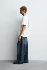 Camiseta blanca de corte boxy fit, cuello redondo y manga corta. Presenta un parche bordado con las letras "NY" y un diseño abstracto en negro en el pecho.