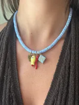 Collar corto con cuentas planas de cerámica celeste claro, con dijes colgantes: hoja dorada, figura amarilla con ojo, chile rojo y caracola celeste.