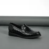Mocasín de cuero vacuno negro con textura grabada tipo reptil. Presenta un diseño clásico con antifaz en el empeine, suela de goma en tono natural y taco bajo de madera.