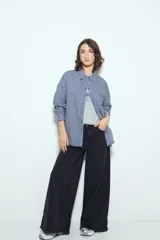 Camisa de silueta amplia y caída fluida, color celeste con rayas verticales blancas. Presenta cuello camisero clásico, cierre frontal con botones y un bolsillo aplicado en el pecho. Las costuras están resaltadas en azul oscuro.