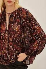 Camisa de corte holgado con estampado ikat en tonos bordó, negro y destellos metalizados. Presenta escote en V con cordones ajustables, mangas largas abullonadas y puños elásticos.