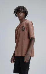 Remera manga corta color marrón (mocca) con un gran estampado trasero en negro que presenta tres máscaras o calaveras estilizadas con una corona en la parte superior y gotas o lágrimas cayendo.