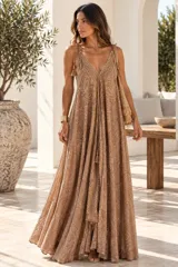 Vestido largo color dorado, confeccionado en tela semitransparente con diseño bordado floral y detalles de brillo. Presenta escote en V, tirantes finos ajustables con borlas en los hombros, y corte amplio y fluido con gran movimiento.