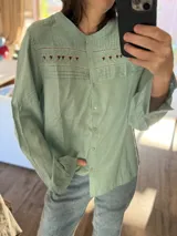 Camisa de manga larga en color verde menta, con cuello mao y cierre frontal de botones. Presenta un diseño con alforzas en el pecho y una franja horizontal con bordados florales y detalles de punto smock. Las mangas tienen puños con volados.