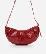 Cartera tipo shoulder bag de diseño media luna, confeccionada en poliuretano con acabado brillante. Presenta detalles frontales estilo gota, cierre superior por cremallera y correa de hombro ajustable con ojetes metálicos.