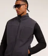 Chaleco deportivo gris oscuro con cuello alto y cierre frontal con cremallera. Presenta recortes en tejido liso en la parte superior del pecho y en los bolsillos laterales.