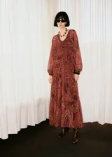 Vestido largo de corte evasé y diseño oversize, confeccionado en tejido semitransparente con estampado paisley en tonos rojizos y marrones. Presenta cuello escotado con cierre frontal, mangas largas con puños abullonados y forro interior a tono.