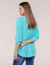Blusa turquesa de silueta suelta confeccionada en tejido liviano, con escote en V, pequeña cartera frontal con botones decorativos y mangas 3/4 con presilla para arremangar. Presenta patrón estampado geométrico tipo tie-dye y ruedo curvo.