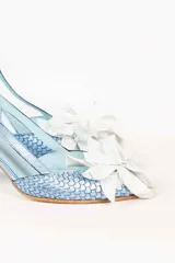 Zapato de tacón celeste con textura tejida, tira trasera y aplique de flor blanca en la punta.