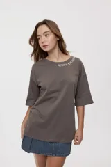 Remera de manga corta y cuello redondo, de corte oversized en color marrón, con texto bordado o estampado a lo largo del escote en el hombro izquierdo.