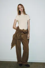 Camiseta de manga corta color marrón oscuro con textura acanalada, cuello redondo y bajo recto. Presenta un bajo superpuesto de tul o tela más clara, visible en el ruedo y en el cuello.