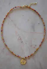 Collar de cristales facetados en tonos naranja, rojo y beige, con esferas doradas intercaladas. Incluye un dije redondo de baño de oro con una cruz de circonitas incrustadas.