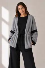 Cárdigan de punto en color gris con detalles en negro en los bordes, puños y una franja horizontal en las mangas. Presenta un diseño abierto con cordón ajustable en la cintura y bolsillos laterales.