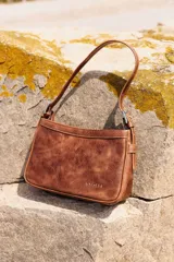 Cartera de hombro compacta de diseño estructurado, confeccionada en cuero vacuno marrón con efecto gastado. Presenta cierre superior y correa de hombro regulable con hebillas metálicas. Cuenta con el logotipo de la marca grabado en la parte frontal inferior.