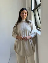Poncho de tejido de punto color beige con cuello alto y diseño de trenzas verticales en el frente. Presenta mangas integradas con puños acanalados y un corte holgado con ruedo redondeado.