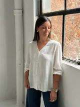Blusa blanca de manga larga con cuello en V y cierre frontal de botones. Presenta una textura sutil en la tela y un corte recto y holgado.