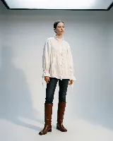 Camisa de algodón blanca con diseño holgado, cuello alto con volados, frente con tablas verticales y cierre frontal con botones. Presenta mangas largas amplias con puños ajustables mediante lazos.