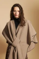 Poncho de tejido suave color beige con diseño envolvente y terminaciones con costuras decorativas en contraste color negro. Presenta un detalle de lazo frontal integrado.