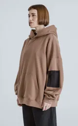 Hoodie oversize color marrón, confeccionado en algodón rústico. Presenta capucha forrada, bolsillo frontal tipo canguro y un bolsillo utilitario con cierre en la manga izquierda, con detalles en microfibra.
