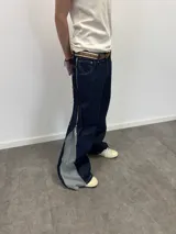 Pantalón de jean azul raw con diseño de corte versátil, ajustable mediante cierres laterales que revelan un panel interior rojo. Presenta una estampa gráfica de gran tamaño en la parte trasera con detalles bordados, costuras reforzadas y etiqueta de cuero en la cintura.