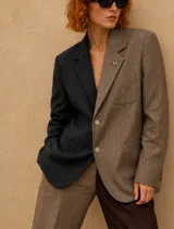 Blazer oversize de diseño bicolor con estructura asimétrica, combinando un lado en color negro y otro en color marrón, ambos con estampado de rayas diplomáticas. Presenta solapas clásicas, cierre frontal con botones de strass y un pin decorativo en forma de perro salchicha.
