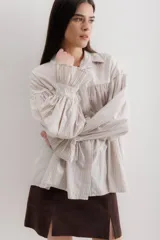 Camisa beige a rayas verticales, de corte holgado, con cuello camisero y mangas largas abullonadas con puños elásticos.