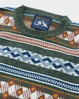 Buzo tejido de punto con diseño de patrones geométricos y rombos en tonos verde, naranja, blanco y azul. Presenta cuello redondo, puños y ruedo acanalados, con etiqueta de marca en el pecho.
