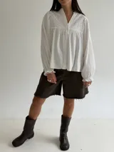Blusa blanca de corte holgado con cuello mao, botones frontales y detalles bordados en el pecho. Presenta mangas largas con puños con volados.