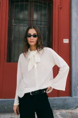 Blusa de lino color crudo, con cuello alto y lazo decorativo frontal. Presenta mangas largas con corte acampanado y diseño entallado con pinzas delanteras.