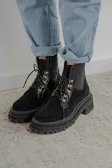 Borcego de gamuza color chocolate con paneles laterales elásticos negros y suela track dentada. Presenta sistema de cordones con ganchos metálicos y tirador en la parte trasera.