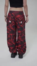 Pantalón de tiro bajo con estampado de camuflaje en tonos rojos y negros. Presenta diseño holgado, detalles de estrellas blancas aplicadas, paneles laterales con franjas verticales y bolsillos cargo.
