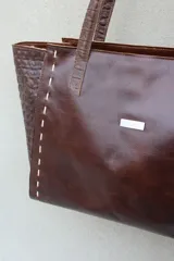 Bolso tipo shopper de gran tamaño confeccionado en cuero vacuno color marrón oscuro. Presenta laterales con textura grabada tipo cocodrilo y detalles de costuras decorativas con tientos de cuero en los paneles frontales y traseros. Incluye doble asa de hombro, cierre superior y placa metálica decorativa en el frente.