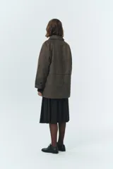 Chaqueta de gamuza sintética color verde militar, con cuello camisero y cierre frontal de botones. Presenta un diseño de corte amplio con bolsillos delanteros de solapa y mangas largas. El material tipo suede ofrece una textura suave y un estilo clásico de inspiración masculina.