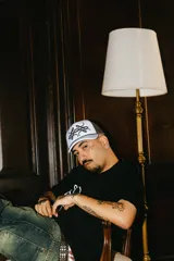 Gorra trucker gris con paneles frontales blancos y paneles traseros de rejilla negra. Presenta un logo bordado en el panel frontal y una cuerda decorativa en la base de la corona.