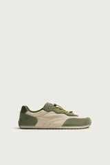 Zapatilla deportiva barefoot fabricada en combinación de piel y tejido, en tonos verde oliva y beige, con cordones elásticos amarillos y suela de goma flexible.