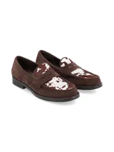 Mocasines de gamuza marrón con detalle de cuero vacuno con estampado de manchas en la parte superior. Presentan una banda frontal característica de los mocasines tipo penny y suela plana de goma.