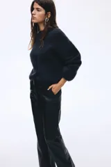 Pantalón de cuero sintético negro con corte bootcut, cierre frontal con botón oculto y costuras decorativas sobre las rodillas.