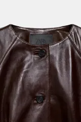 Chaqueta corta de piel color marrón, con cuello redondo, manga larga y cierre frontal con botones. Presenta un diseño con elástico en el ruedo y forro interior.
