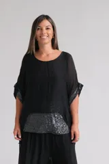 Blusa negra de corte holgado y diseño asimétrico, confeccionada en tela semitransparente. Presenta detalles de lentejuelas brillantes en el ruedo inferior y en los bordes de las mangas cortas.