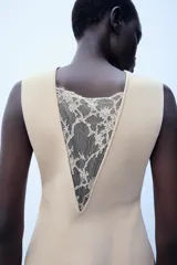 Vestido midi de color beige, con cuello redondo y diseño sin mangas. Presenta un cuerpo liso de punto con bajo asimétrico y una falda inferior confeccionada en encaje tono sobre tono.