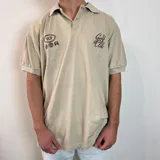 Chomba color beige con cuello clásico, cierre de dos botones y mangas cortas. Presenta bordados en el frente con las inscripciones "VCP" y "Golf Club". Corte oversize.