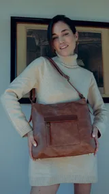 Cartera marrón de simil cuero con bolsillo frontal con tapa y cierre dorado. Tiene correa larga regulable y asa de mano.