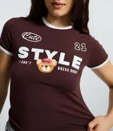 Remera de corte ajustado color marrón chocolate, con cuello redondo y bordes de mangas en color blanco. Presenta un estampado frontal con la palabra 'STYLE' en letras grandes, un oso pequeño con gorra y el texto 'Cute' y '21'.